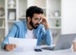 Stressed young man looking at laptop screen and holding documents