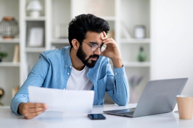 Stressed young man looking at laptop screen and holding documents