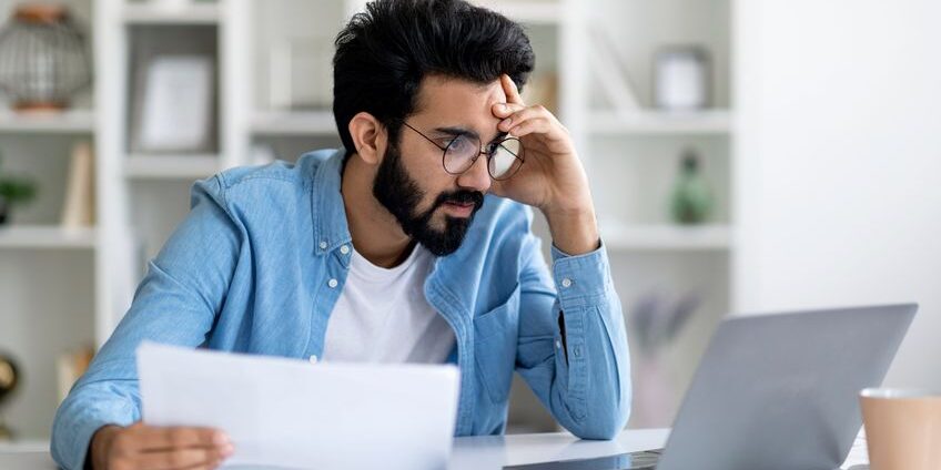 Stressed young man looking at laptop screen and holding documents